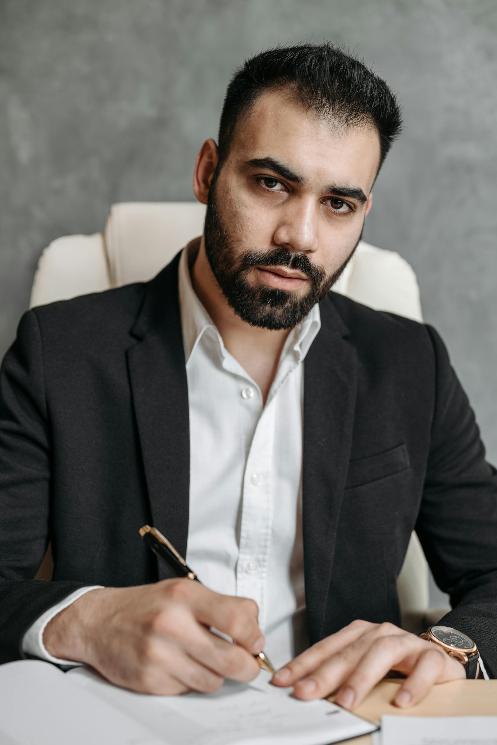Serious man in a black suit signing documents in an office setting.