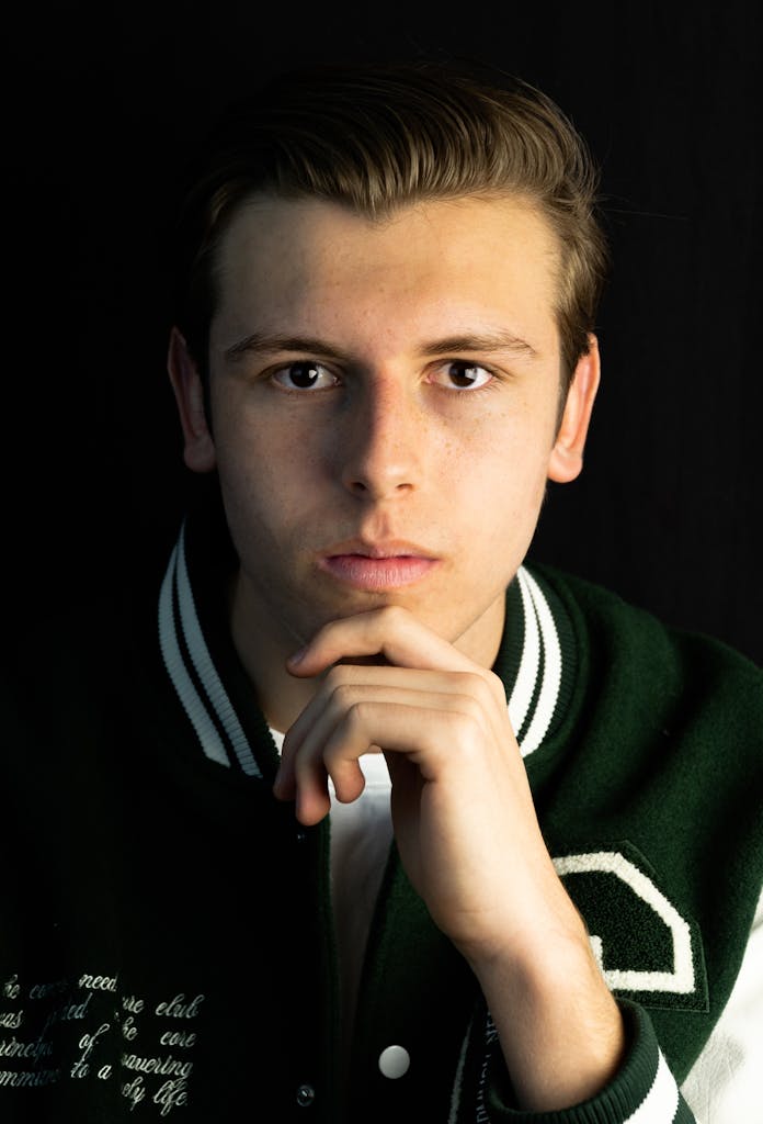 Close-up portrait of a young man in a green varsity jacket against a black background.